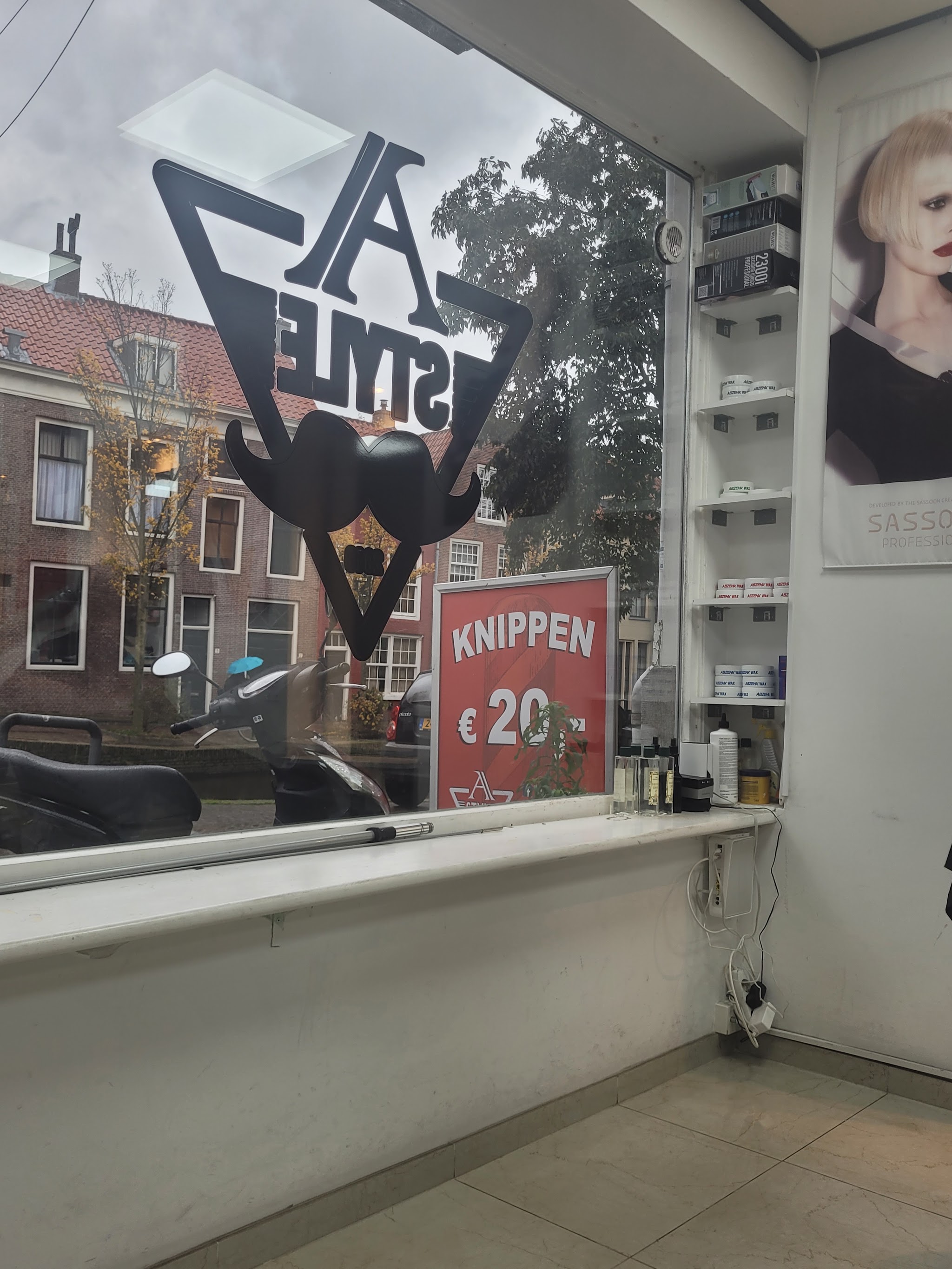 Het raam van A Style van binnenuit gezien, met het rode bord Knippen 20 euro en de huizen aan de overkant